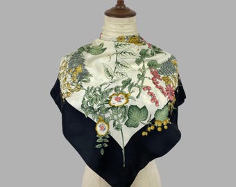 May include: A square scarf featuring a cream-colored center with a botanical print of green leaves, yellow flowers, and clusters of red and yellow berries. The scarf has a wide, solid black border along the bottom edges and is draped on a mannequin.