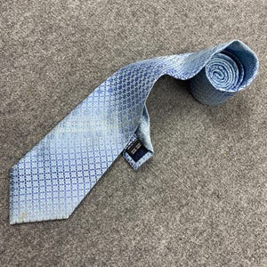 Puede incluir: Una corbata azul claro con un patrón geométrico. La corbata está enrollada en el extremo, mostrando el intrincado diseño. La corbata está hecha de un material brillante y está sobre una superficie gris.
