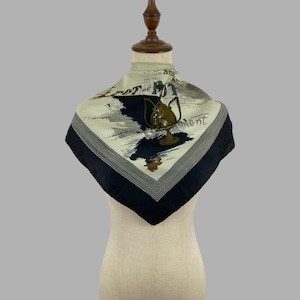 Pode incluir: Um lenço quadrado com centro na cor creme, borda azul-escura e um gráfico central de um bule dourado ornamentado. O design inclui texto preto em cursivo e formas abstratas azul-escuras, emoldurado por linhas finas na cor cinza.