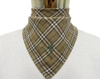 Può includere: Un foulard triangolare con un motivo a quadri nei colori beige, marrone, bianco e rosso. Il tessuto è ricamato con la parola Burberry e un piccolo logo a forma di cavaliere in filo verde. Il foulard è drappeggiato su un supporto cilindrico.