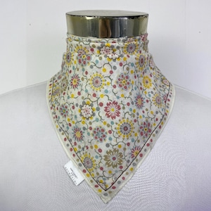 May include: A floral patterned neck scarf in a triangular shape. The scarf features a white background with a repeating pattern of flowers in shades of pink, yellow, and blue. The edges are bordered with a dotted pattern. The tag reads "INTERMODE".