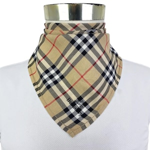 May include: A beige bandana featuring a classic plaid pattern in black, white, and red. The fabric is folded into a triangular shape around a neck form. The word Burberry is printed in black on the lower section of the fabric, along with a logo.