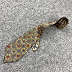 Pode incluir: Uma gravata cinza com um padrão repetido de desenhos florais vermelhos e dourados. A gravata está enrolada na extremidade e tem uma pequena etiqueta anexada. A gravata está sobre uma superfície cinza.