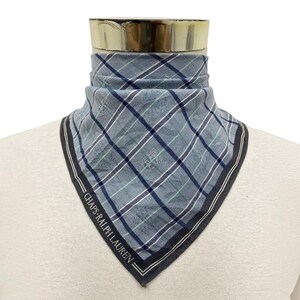 May include: A triangular neck scarf featuring a light blue, navy, and white plaid pattern with thin green accent lines. The fabric displays a repeating crest logo and the text CHAPS RALPH LAUREN printed along the dark navy border edge.