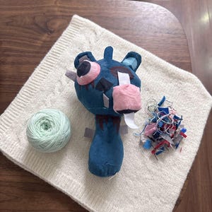 Op de afbeelding: Een donkerblauw pluche speelgoed met een groot roze oog, een witte rechthoekige lap over het andere oog en een roze snuit. Naast het speelgoed ligt een bol lichtgroen garen en een kleine hoop diverse gekleurde stofresten en losse draden.