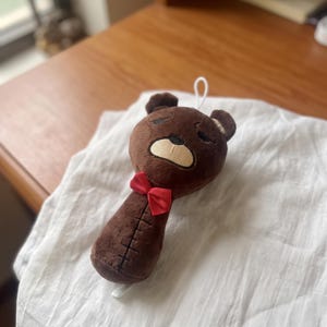 Op de afbeelding: Een bruin pluche speeltje in de vorm van een berenkop met een lange nek, voorzien van een kleine rode strik bij de nek, geborduurde gelaatstrekken met gesloten ogen en een verticale stiklijn die over het midden van het onderste lichaamsdeel loopt.