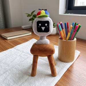 Puede incluir: Un peluche que representa un personaje de cabeza cuadrada gris con una cara de pantalla negra, ojos circulares blancos y una nariz triangular. El personaje lleva una pequeña gorra multicolor con paneles rojos, amarillos, azules y verdes sobre un taburete marrón.
