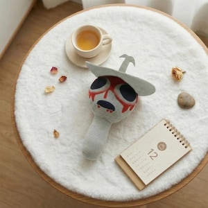 Puede incluir: Un peluche con cuerpo gris, cabeza redonda con grandes ojos negros y diseño rojo goteante. Tiene un sombrero gris con detalle de espada. Un pequeño calendario y una taza de té también están sobre la superficie blanca y esponjosa.