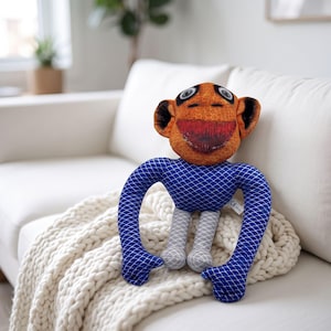May include: A plush toy featuring an orange, textured head with large eyes and a wide, dark red mouth. The body is covered in a blue fabric with a white scale pattern, while the legs are light gray with a white diamond grid design.