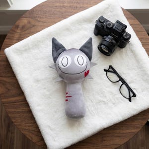 Può includere: Un giocattolo a forma di gatto di peluche grigio con grandi occhi, una bocca sorridente e un dettaglio a cuore rosso. Il giocattolo ha orecchie grigie e un colletto a punte. Una fotocamera nera e occhiali da vista poggiano su una superficie bianca e soffice.