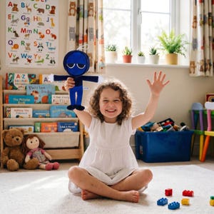 Könnte beinhalten: Ein lächelndes Mädchen hält eine blaue Plüschpuppe mit Kapuze. Die Puppe hat eine weiße Schnur befestigt. Das Mädchen sitzt auf einem weißen Teppich in einem Raum mit einem Bücherregal, Spielzeug und einem Fenster.