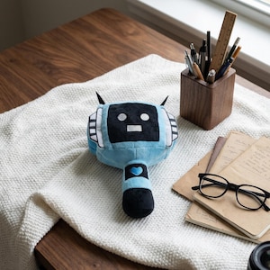 Pode incluir: Um brinquedo de pelúcia azul em forma de robô com um rosto preto e antenas. O robô tem um design de coração em sua haste. Um porta-lápis de madeira com canetas e lápis, cadernos e óculos também estão na imagem.