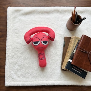 May include: A red plush toy with a cartoon face and a phone-like shape. The toy has black-rimmed eyes and a small mouth. It is lying on a white, fluffy blanket. A wooden pencil holder and books are in the background.