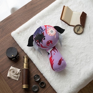 May include: A plush zombie doll with a purple head and body, black ears, and a white string for hanging. The doll has a bloodied eye and red accents. A compass, journal, and telescope are nearby.
