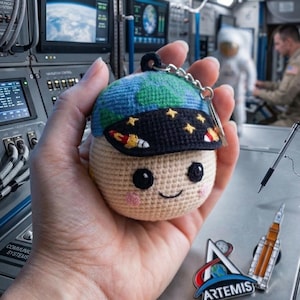 May include: A crocheted keychain depicting a smiling character wearing a blue and green Earth-themed hat and a black visor with stars. The keychain is held in a hand, with space-themed pins and a pen in the background.