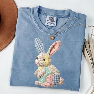 May include: A light blue Comfort Colors t-shirt with a patchwork bunny design. The bunny features floral, gingham, and polka dot patterns in pastel colors. A gold necklace with a pendant rests on the shirt. A brown hat and dried plant are in the background.