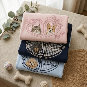 May include: Three folded sweatshirts in light pink, navy blue, and light blue. Each features embroidered heart-shaped frames with gingham patterns, portraits of cats and dogs, and the names Luna, Archie, Bruno, and Daisy stitched in cursive.