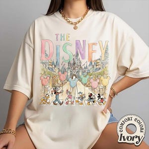 K&ouml;nnte beinhalten: Elfenbeinfarbenes T-Shirt mit einer farbenfrohen Illustration von Disney-Figuren, die eine Stra&szlig;e vor einem Schloss &uuml;berqueren. Der Text "THE DISNEY" befindet sich &uuml;ber der Illustration. Das Shirt hat ein Comfort Colors-Label.