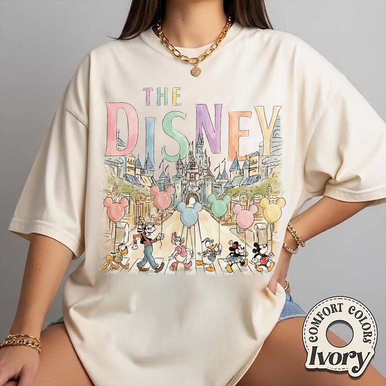 K&ouml;nnte beinhalten: Elfenbeinfarbenes T-Shirt mit einer farbenfrohen Illustration von Disney-Figuren, die eine Stra&szlig;e vor einem Schloss &uuml;berqueren. Der Text "THE DISNEY" befindet sich &uuml;ber der Illustration. Das Shirt hat ein Comfort Colors-Label.