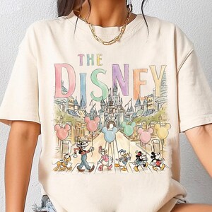 K&ouml;nnte beinhalten: Cremefarbenes T-Shirt mit einer bunten Grafik von Disney-Figuren, die eine Stra&szlig;e vor einem Schloss &uuml;berqueren. Das Wort "DISNEY" wird in Pastellfarben &uuml;ber der Szene mit Mickey Mouse-Ballons angezeigt.