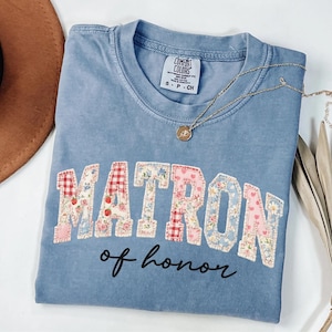 Puede incluir: Camiseta azul claro con la frase "MATRON of honor" en un diseño de patchwork. Las letras están decoradas con estampados florales y cuadros. Un collar dorado con un colgante está sobre la camiseta.