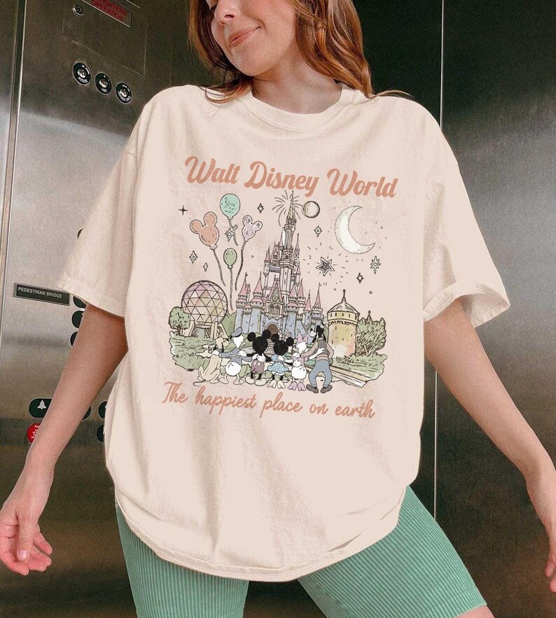 May include: A cream-coloured t-shirt with a graphic of Cinderella's castle, Mickey Mouse, and other Disney characters. The text "Walt Disney World" and "The happiest place on earth" are printed above and below the graphic in a vintage style.