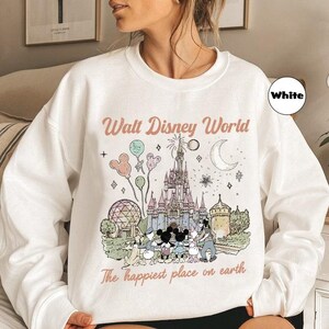 May include: White sweatshirt with a graphic of Cinderella's castle, Mickey Mouse, and other Disney characters. The text "Walt Disney World" and "The happiest place on earth" are printed on the front.