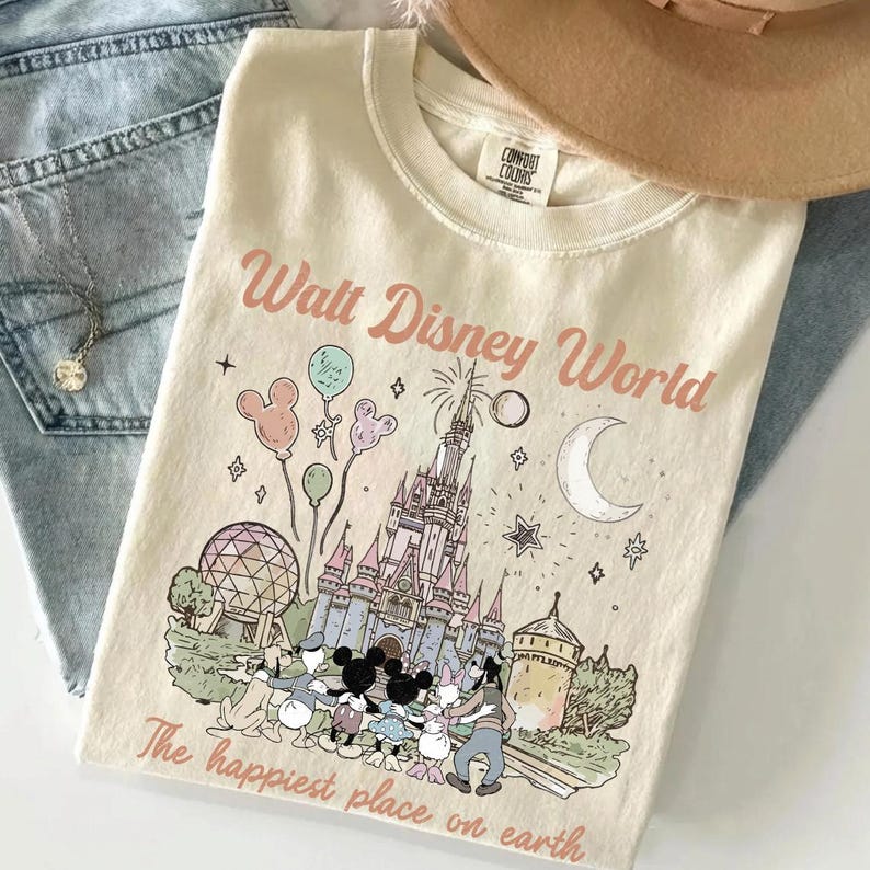 May include: Cream-coloured t-shirt featuring a graphic of the Walt Disney World castle, Mickey Mouse, Minnie Mouse, and other characters. The text "Walt Disney World" and "The happiest place on earth" are printed on the shirt.