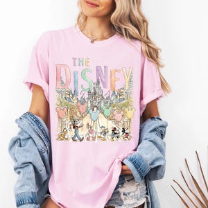 K&ouml;nnte beinhalten: Hellrosa T-Shirt mit dem Text "THE DISNEY" in Pastellfarben. Darunter eine Aquarellillustration eines Schlosses und Disney-Figuren. Das T-Shirt hat kurze &Auml;rmel und einen Rundhalsausschnitt.