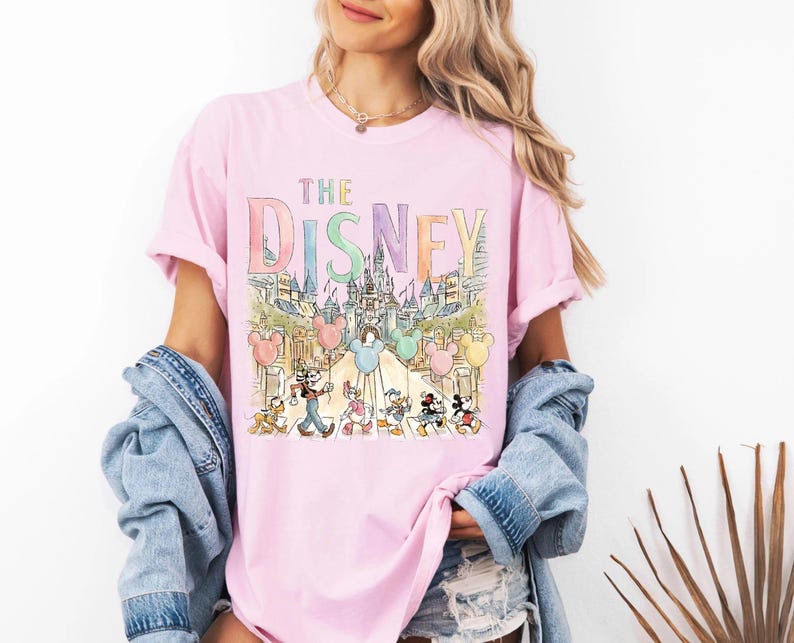 K&ouml;nnte beinhalten: Hellrosa T-Shirt mit dem Text "THE DISNEY" in Pastellfarben. Darunter eine Aquarellillustration eines Schlosses und Disney-Figuren. Das T-Shirt hat kurze &Auml;rmel und einen Rundhalsausschnitt.