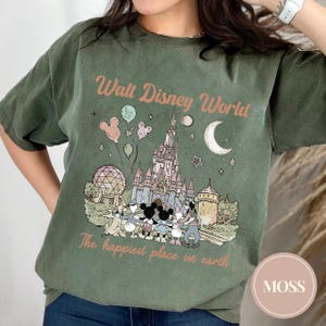 May include: An olive green t-shirt featuring a graphic of Cinderella's castle, Mickey Mouse, and other Disney characters. The text "Walt Disney World" and "The happiest place on earth" are printed on the shirt.
