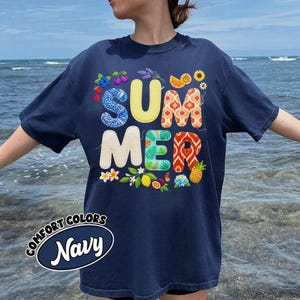 以下が含まれることがあります： ネイビーブルーのComfort Colors Tシャツ。カラフルで模様入りの文字で「SUMMER」と書かれています。デザインには花や果物のイラストが含まれています。ゆったりとしたフィット感のTシャツです。