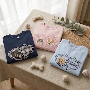 May include: Three crewneck sweatshirts in navy blue, light pink, and light blue. Each features an embroidered heart-shaped locket design with a gingham pattern, a pet portrait, and a name: Bruno, Luna, Archie, or Bella in cursive script.