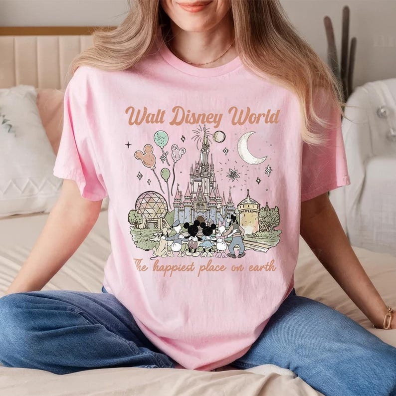 May include: Pale pink t-shirt featuring a Disney World graphic with Cinderella's castle, Mickey Mouse, and other characters. The text reads "Walt Disney World" and "The happiest place on earth".
