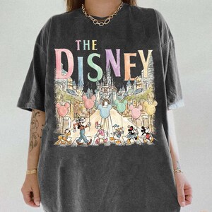 K&ouml;nnte beinhalten: Dunkelgraues T-Shirt mit dem Text "THE DISNEY" in Pastellfarben, &uuml;ber einer farbenfrohen Illustration eines Schlosses, Mickey Mouse-Ballons und Disney-Figuren. Das Shirt hat kurze &Auml;rmel und eine lockere Passform.