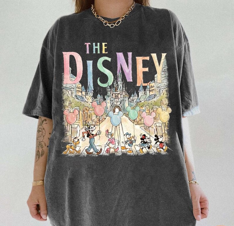 K&ouml;nnte beinhalten: Dunkelgraues T-Shirt mit dem Text "THE DISNEY" in Pastellfarben, &uuml;ber einer farbenfrohen Illustration eines Schlosses, Mickey Mouse-Ballons und Disney-Figuren. Das Shirt hat kurze &Auml;rmel und eine lockere Passform.