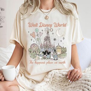 May include: Cream-coloured t-shirt with a pastel illustration of Cinderella's castle, Mickey Mouse, and other Disney characters. The text "Walt Disney World" and "The happiest place on earth" are printed above and below the graphic.