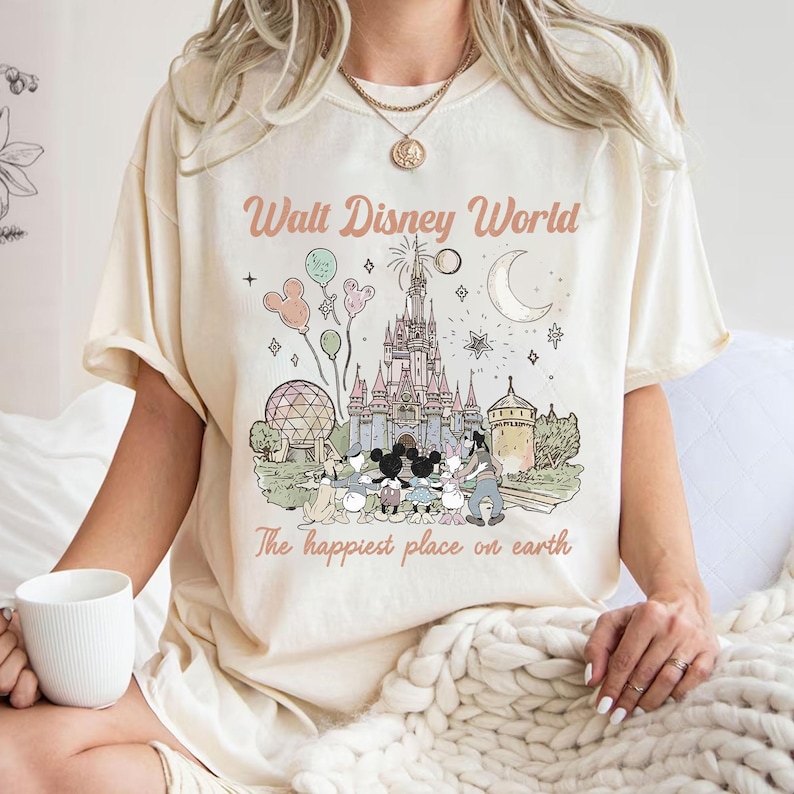 May include: Cream-coloured t-shirt with a pastel illustration of Cinderella's castle, Mickey Mouse, and other Disney characters. The text "Walt Disney World" and "The happiest place on earth" are printed above and below the graphic.