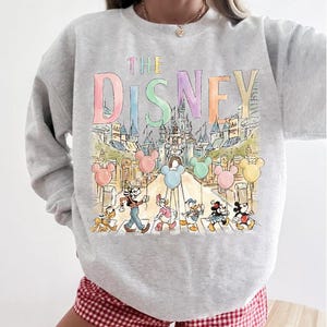 K&ouml;nnte beinhalten: Hellgraues Sweatshirt mit einer farbenfrohen Illustration von Disney-Figuren, die eine Stra&szlig;e &uuml;berqueren, mit Mickey Mouse-Ballons. Die Worte "THE DISNEY" sind oben in Pastellfarben aufgedruckt. Das Sweatshirt wird mit rot-wei&szlig; karierten Shorts getragen.