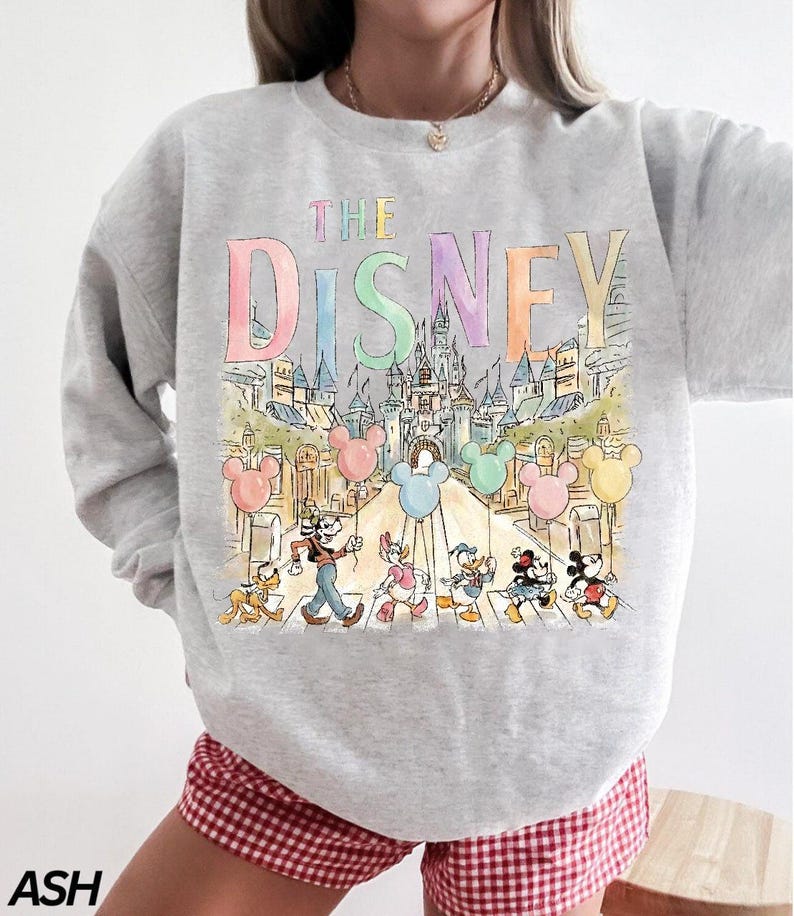 K&ouml;nnte beinhalten: Hellgraues Sweatshirt mit einer farbenfrohen Illustration von Disney-Figuren, die eine Stra&szlig;e &uuml;berqueren, mit Mickey Mouse-Ballons. Die Worte "THE DISNEY" sind oben in Pastellfarben aufgedruckt. Das Sweatshirt wird mit rot-wei&szlig; karierten Shorts getragen.