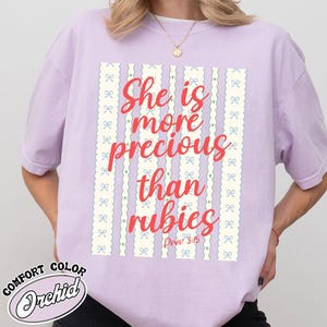 Puede incluir: Camiseta de color lila con el texto "She is more precious than rubies" en escritura roja. El diseño incluye rayas verticales con lazos y corazones. La camiseta está etiquetada como "Comfort Color Orchid".