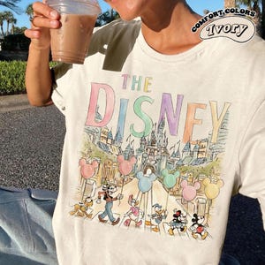 K&ouml;nnte beinhalten: Elfenbeinfarbenes Comfort Colors T-Shirt mit einer farbenfrohen Illustration von Disney-Figuren und dem Text "THE DISNEY". Das Design umfasst Micky Maus, Minnie Maus, Goofy, Donald Duck und Daisy Duck, mit einem Schloss und Ballons.