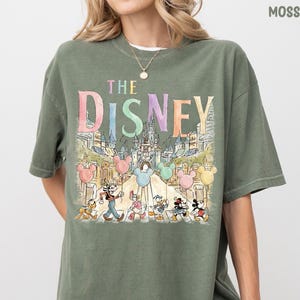 K&ouml;nnte beinhalten: Moosgr&uuml;nes T-Shirt mit einer farbenfrohen Grafik von Disney-Figuren und dem Text "THE DISNEY". Das Design umfasst Micky Maus, Donald Duck und andere Charaktere mit einer Schlosskulisse und Mickey Mouse-Ballons.