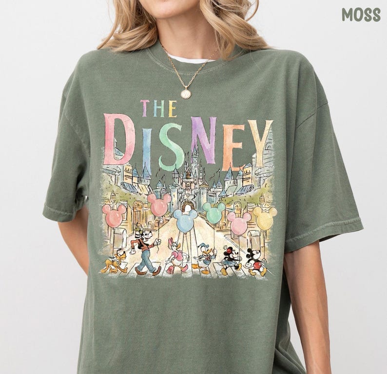 K&ouml;nnte beinhalten: Moosgr&uuml;nes T-Shirt mit einer farbenfrohen Grafik von Disney-Figuren und dem Text "THE DISNEY". Das Design umfasst Micky Maus, Donald Duck und andere Charaktere mit einer Schlosskulisse und Mickey Mouse-Ballons.