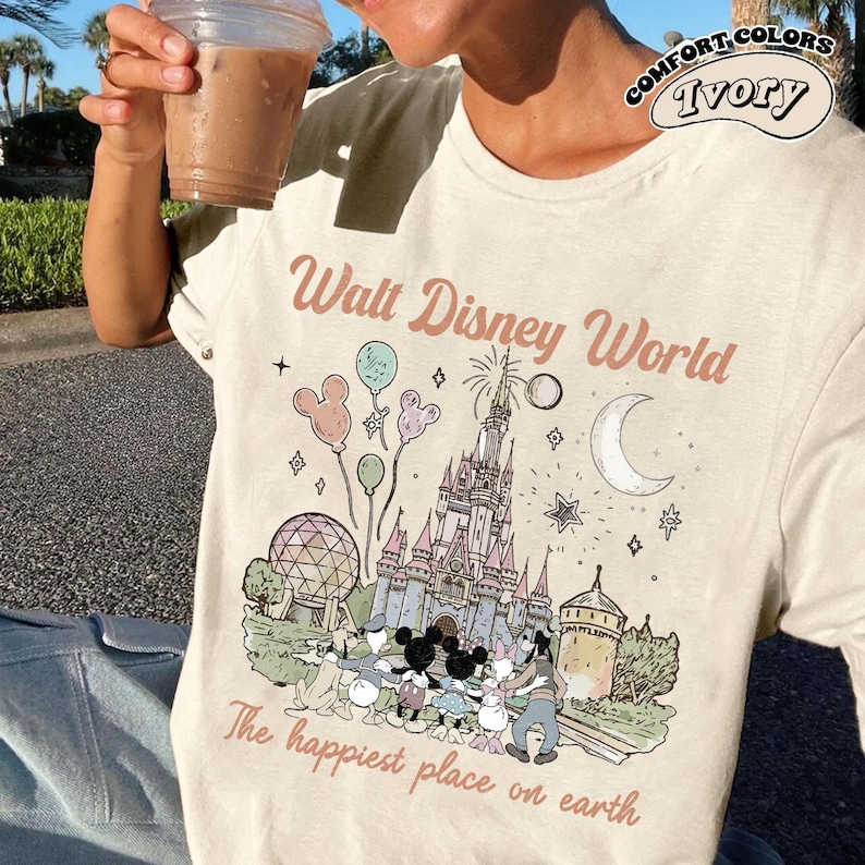 May include: Ivory Comfort Colors t-shirt with a Walt Disney World graphic. The design includes a castle, Mickey Mouse and friends, balloons, and the text "The happiest place on earth."