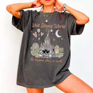May include: Dark grey t-shirt with a vintage-style Walt Disney World graphic. The design features the castle, Mickey Mouse and friends, balloons, and the text "The happiest place on earth."