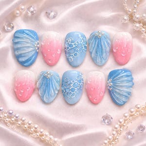 May include: A collection of ten artificial nails with various designs. The nails feature a gradient of pink and white, blue with bubble patterns, and blue with shell-like designs. Some nails have small, pearl-like embellishments. The background is a satin fabric.