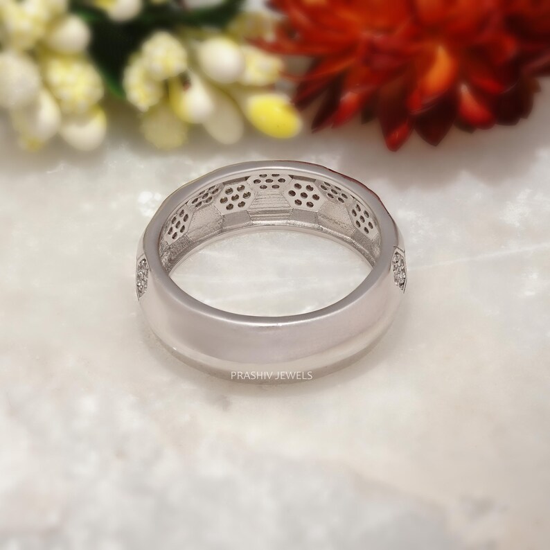 May include: A silver ring with a geometric design and small, clear stones. The ring has a polished finish and the brand name "PRASHIV JEWELS" is visible. The ring is set against a blurred background of flowers.