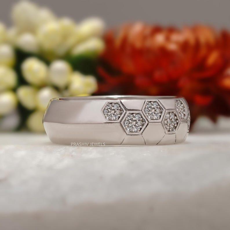 May include: A silver ring with a geometric honeycomb design. The ring features a series of hexagonal shapes, each filled with small, sparkling stones. The ring is set against a blurred background of flowers.