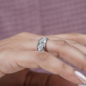 May include: A silver ring with a honeycomb design, set with small, sparkling stones. The ring is on a finger, with the brand name "PRASHIN JEWELS" visible. The ring's design is a series of connected hexagons, each filled with a cluster of small stones.